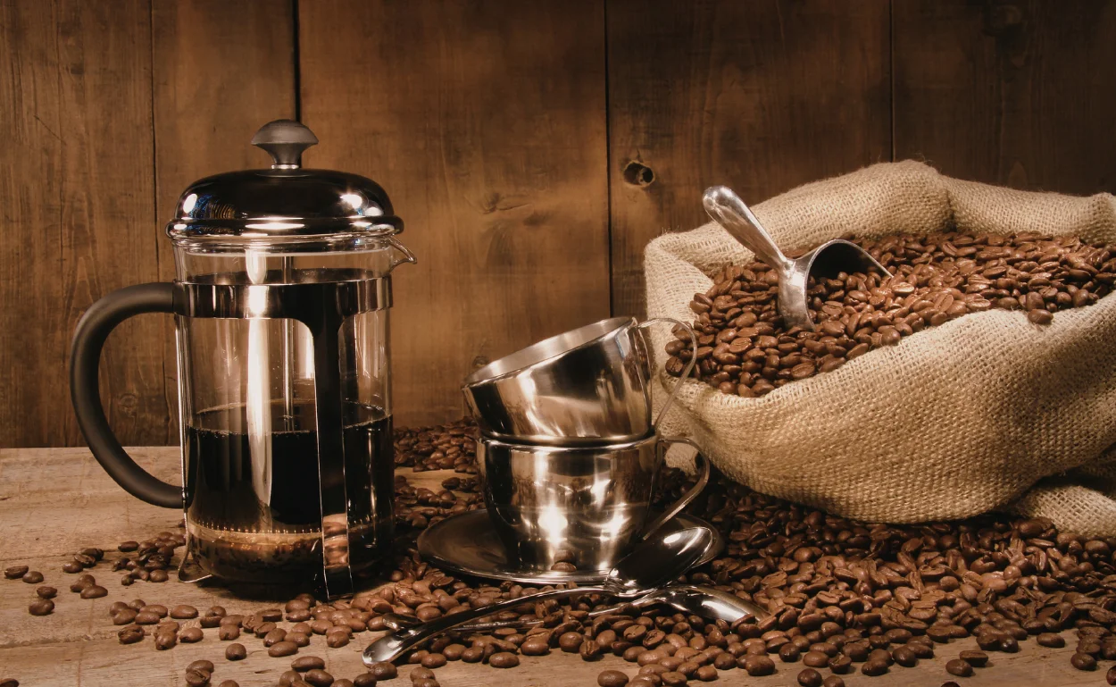 French press coffee maker with whole coffee beans in a burlap sack and metal cups